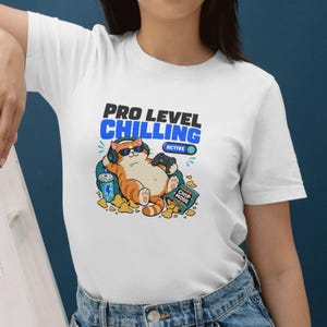 Puede incluir: Camiseta blanca con un gato de dibujos animados con gafas de sol y auriculares, sosteniendo un mando de videojuegos. La camiseta tiene el texto "PRO LEVEL CHILLING ACTIVE" encima del gato, con una bebida y una bolsa de patatas fritas. El gato es naranja y blanco.