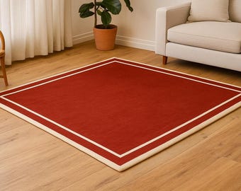 May include: A square, red rug with a cream-colored border and a double cream-colored inner border. The rug is placed on a hardwood floor. The rug is a home decor item.