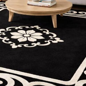 May include: A black area rug with a white floral design in the center and a white border with a scrolling pattern. The rug is rectangular and sits on a hardwood floor. A round wooden coffee table is in the background.