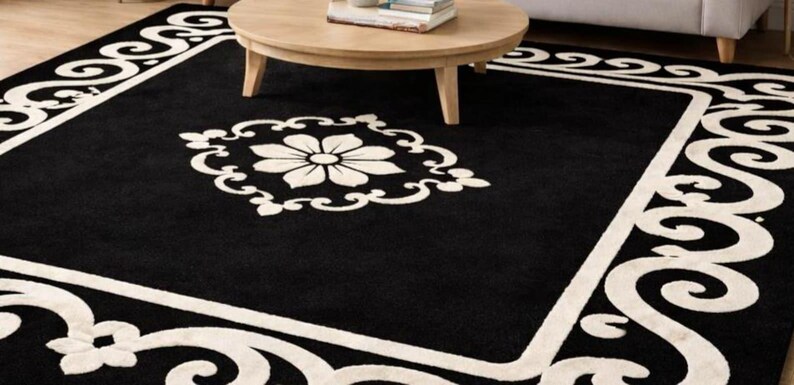 May include: A black area rug with a white floral design in the center and a white border with a scrolling pattern. The rug is rectangular and sits on a hardwood floor. A round wooden coffee table is in the background.