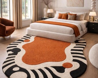 May include: An area rug with an irregular, organic shape featuring a large central burnt orange section surrounded by cream-colored areas and black-and-white striped borders. The rug includes circular orange and black accents throughout the design.
