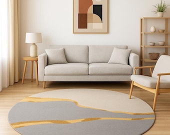 May include: A round area rug with a modern design. The rug features a gray base with a light beige upper section and gold-colored accent lines. The rug is placed in a living room setting with a sofa, artwork, and a side table.