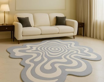May include: A unique, abstract-shaped rug with a light gray and off-white wavy pattern. The rug is placed on a light-colored floor in front of a cream-colored sofa with dark gray throw pillows. The room has a neutral color palette.