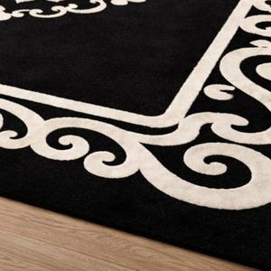 May include: A black rectangular rug with a cream-colored ornate border and central design. The rug's design features scrolling patterns and floral elements, creating a classic and elegant aesthetic. The rug is placed on a light-colored wooden floor.