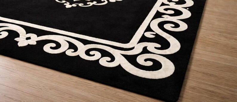 May include: A black rectangular rug with a cream-colored ornate border and central design. The rug's design features scrolling patterns and floral elements, creating a classic and elegant aesthetic. The rug is placed on a light-colored wooden floor.
