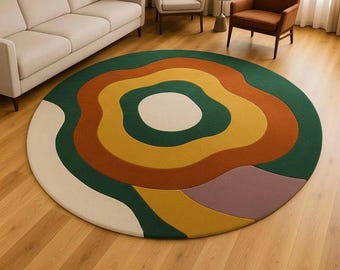 May include: A round area rug with a colorful, abstract design. The rug features concentric rings of green, orange, yellow, and purple, with a white central shape. The rug is placed on a hardwood floor.