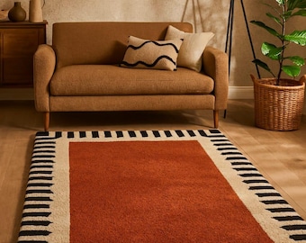 May include: A rectangular rug with a burnt orange center and a border of black and cream-colored stripes. The rug is placed on a wooden floor in a living room setting, with a brown sofa and decorative pillows in the background.