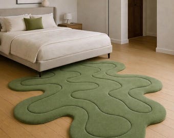 May include: A green, organic-shaped rug on a hardwood floor in a bedroom setting. The rug's design features flowing, irregular lines and a soft, textured surface. The bedroom includes a bed with white linens and a neutral color palette.