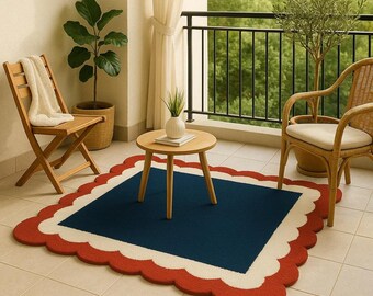 May include: A navy blue rug with a scalloped edge in red and white sits on a tiled patio. A small wooden table and two wicker chairs are arranged around the rug. A folded wooden chair and potted plants complete the outdoor scene.