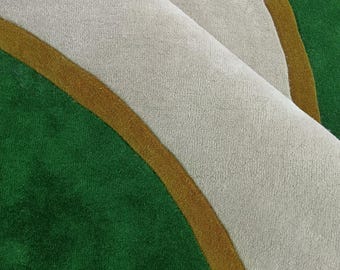 May include: A tufted rug featuring a bold geometric pattern with large sections of deep green and light gray, separated by a thin, curved mustard yellow border. The rug is folded over, revealing the dense, soft texture of the pile surface.