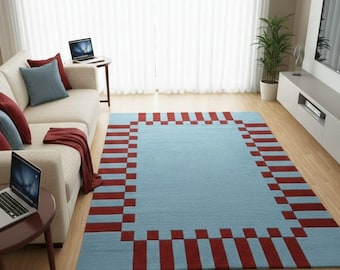 May include: A rectangular area rug with a light blue center and a red and blue checkered border. The rug is placed on a hardwood floor in a living room setting. A beige sofa with pillows and a side table with a laptop are visible.