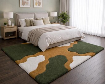 May include: A rectangular area rug featuring a thick, textured pile with an abstract, organic pattern of interlocking shapes in deep forest green, mustard yellow, and off-white, positioned on a dark brown wooden floor in a bedroom.