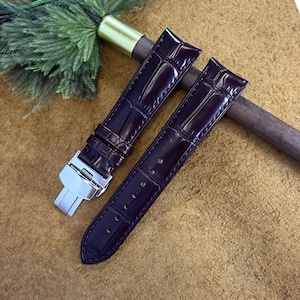 May include: Two dark burgundy watch straps with a crocodile skin effect. The straps feature a silver clasp and are presented on a brown surface. These watch straps are a stylish accessory.