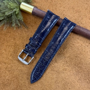 Puede incluir: Dos correas de reloj azul marino con textura de piel de cocodrilo. Una correa tiene una hebilla plateada. Las correas están sobre una superficie marrón con una varilla de madera y una pieza metálica dorada.