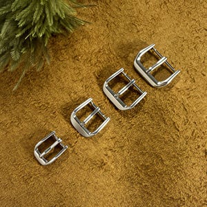 May include: Four silver-colored watch buckles arranged diagonally on a textured, golden-brown surface. Each buckle has a rectangular frame with a central bar and a curved, rounded end. A sprig of green foliage is in the upper left corner.