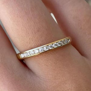 May include: A gold band ring with a row of small, clear gemstones set across the top. The ring is on a finger, and the background is blurred. The ring is likely made of gold and the gemstones are likely diamonds.