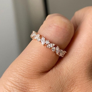 May include: A rose gold ring with a continuous pattern of marquise-cut and round-cut white gemstones. The ring is on a finger, showcasing the intricate design and sparkle of the gemstones. The ring is from NYC Diamond Studio.
