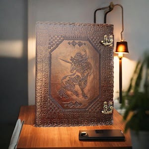 Könnte beinhalten: Ein großes, braunes, ledergebundenes Notizbuch mit einem geprägten Einhorn-Design auf dem Einband. Das Notizbuch hat dekorative Metallverschlüsse und aufwendige Bordürenmuster. Es liegt auf einer Holzoberfläche.