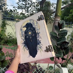 May include: A rectangular journal with a cream-colored cover featuring a black raven illustration centered within a gold circular frame. The raven is adorned with a celestial-style clockwork design on its chest. It has antique brass metal latches.