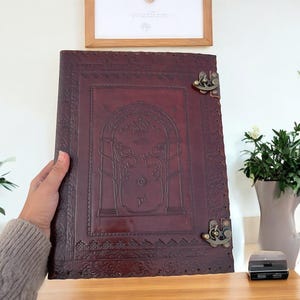 May include: A dark burgundy leather journal with an embossed design featuring an arched gateway, trees, and a crown. The journal has decorative edges and antique brass clasps. The journal is held by a person.