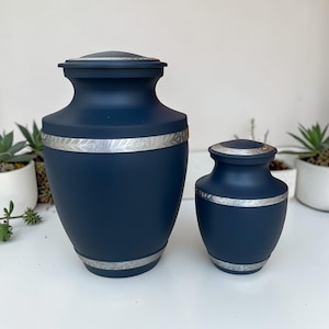 Peut inclure: Deux urnes bleu marine avec des bandes argentées. La plus grande urne a une forme arrondie avec un couvercle, et la plus petite urne a un design similaire. Les deux urnes ont une bande argentée avec un motif floral au milieu.