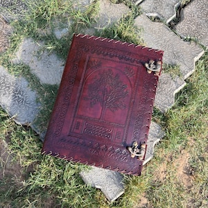 May include: A burgundy leather journal with a tree of life design embossed on the cover. The journal has decorative stitching around the edges and a metal clasp closure. The journal is lying on a bed of grass and stone pavers.