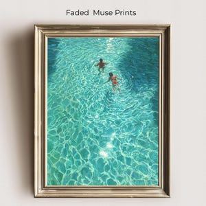 May include: A framed art print titled "Faded Muse Prints" depicts two people swimming in a turquoise pool. The water's surface shimmers with sunlight. The artwork is encased in a gold-toned frame.
