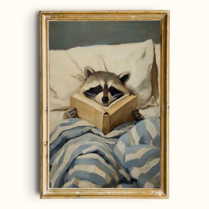 May include: A framed painting shows a raccoon reading in bed. The raccoon is surrounded by pillows and a blue and white striped blanket. The artwork has a vintage, painterly style and is presented in a gold-coloured frame.