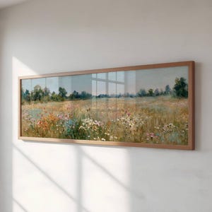 May include: A rectangular framed artwork showing a field of wildflowers in shades of yellow, pink, and white. The painting has a backdrop of trees and a soft blue sky. The frame is made of light brown wood.