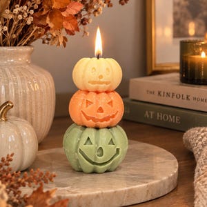 May include: A stacked pillar candle featuring three carved pumpkin shapes in light yellow, orange, and sage green. Each pumpkin has a unique jack-o'-lantern face with cut-out eyes, noses, and mouths. The top pumpkin is lit with a single flame.