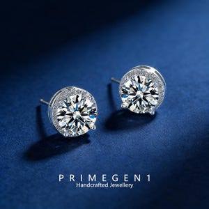 Puede incluir: Un par de pendientes de plata con grandes gemas redondas y transparentes. Cada pendiente está rodeado por un halo de piedras más pequeñas y brillantes. Los pendientes están sobre un fondo azul oscuro. El texto "PRIMEGEN 1 Handcrafted Jewellery" está en la parte inferior.