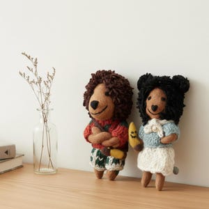 May include: Two handmade bear dolls. One doll wears a red sweater with green accents and green shorts. The other doll wears a light blue sweater and white skirt. Both dolls have brown fur and are holding a yellow banana.