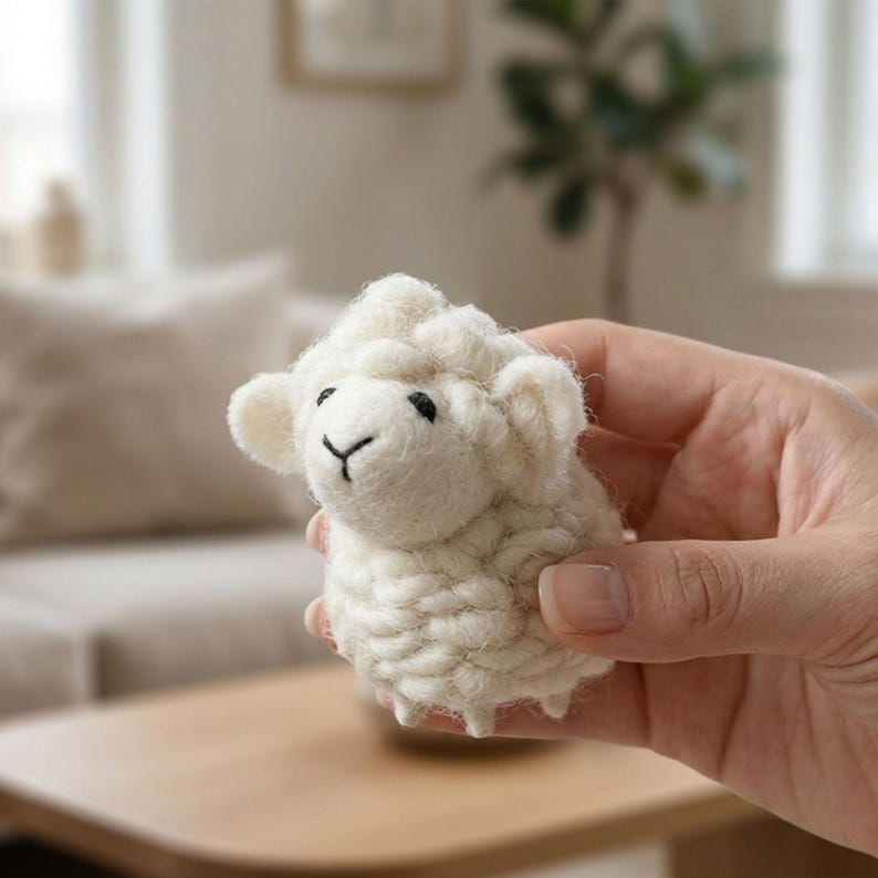 May include: A small, off-white sheep figurine with a fluffy wool texture. The sheep has black eyes and a simple, stitched mouth. The figurine is held in a hand, showcasing its size and detail. This handcrafted item is likely a decorative piece or a child's toy.