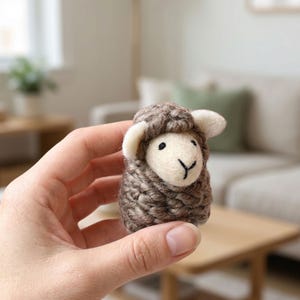 May include: A handcrafted, miniature sheep figurine. The sheep is made of brown and cream-colored wool, with a textured, woven appearance. The sheep has a white face, ears, and a black embroidered face. The figurine is held in a person's hand.