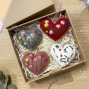 Puede incluir: Un juego de cuatro adornos en forma de corazón en una caja de cartón. Cada corazón de fieltro es de un color diferente: gris, rojo, rosa y blanco. Están decorados con diseños florales bordados y tienen una cuerda para colgar. Una etiqueta dice "To My Love."
