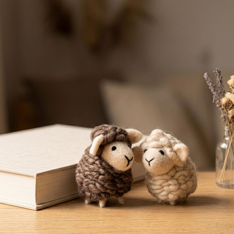 May include: Two handmade wool sheep figurines. One sheep is brown, the other is cream-colored. Both have white faces and small black eyes. They are standing on a wooden surface next to a closed book.