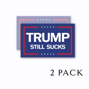 May include: Two blue and red political yard signs with the text "Trump Still Sucks" in white letters. The signs are stacked on top of each other and the text "2 Pack" is printed below the signs.