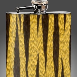 Puede incluir: Petaca de acero inoxidable con un diseño de rayas de tigre en tonos amarillos y marrones. La petaca tiene una tapa y bisagra plateadas. El cuerpo rectangular de la petaca presenta un patrón texturizado. Mide unos 12 cm de alto.