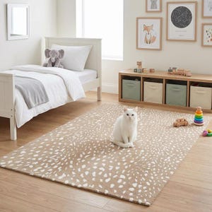 May include: A rectangular area rug featuring a tan background with a repeating white spotted pattern. A white cat sits in the center of the rug, which is placed on a light wood floor in a room with a white bed, a storage unit, and wall art.