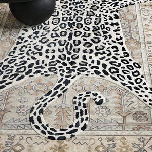 May include: A leopard skin-shaped rug in cream with black spots and a long tail. The rug is laid on a patterned beige rug, creating a layered look. A black round table is partially visible in the upper left corner.