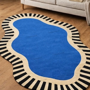 Könnte beinhalten: Ein blauer, freiformiger Teppich mit cremefarbenem Rand und schwarzen Streifen. Der Teppich liegt auf einem Holzboden, im Hintergrund sind ein Sofa und ein Kissen zu sehen. Die einzigartige Form und das Farbschema des Teppichs verleihen dem Raum eine moderne Note.
