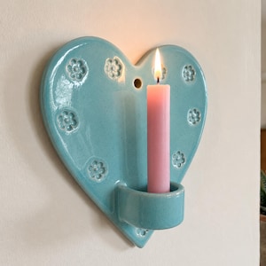 Puede incluir: Un candelabro de pared en forma de corazón con un esmalte azul claro brillante, que presenta patrones de flores en relieve en la superficie y un pequeño orificio circular en la parte superior. Una vela rosa está encendida en el soporte.