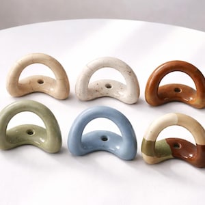 May include: Six ceramic D-shaped drawer pulls in shades of beige, white, brown, green, blue, and a multi-tone design. Each pull has a small hole for mounting. The pulls are displayed on a white surface.
