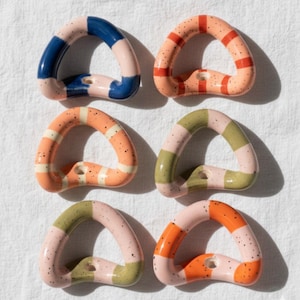 Puede incluir: Seis objetos cerámicos en forma de anillo de colores con superficies moteadas. Cada anillo presenta una combinación de colores única, incluyendo azul y rosa, naranja y rayas rojas, y verde y rosa. Los objetos están dispuestos sobre una superficie blanca.
