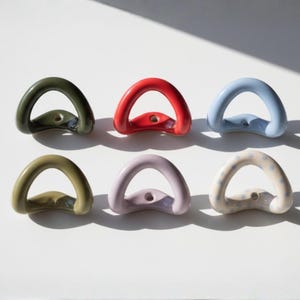 May include: Six ceramic drawer pulls in various colors. The pulls are arched with a circular opening and a hole at the base. Colors include olive green, red, light blue, pale green, lavender, and white with blue polka dots. The pulls are arranged on a white surface.