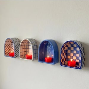 May include: Four ceramic wall-mounted candle holders in a row. Each holder has a different design: polka dots, grid, solid blue, and checkered. Each holds a lit red candle, creating a warm glow.