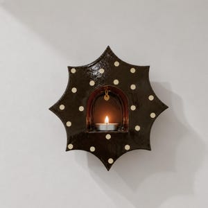 Peut inclure: Un bougeoir mural en forme d'étoile brun foncé avec une finition brillante et un motif de petits pois clairs. Le centre présente une alcôve arquée contenant une bougie chauffe-plat allumée avec un petit anneau de couleur dorée.