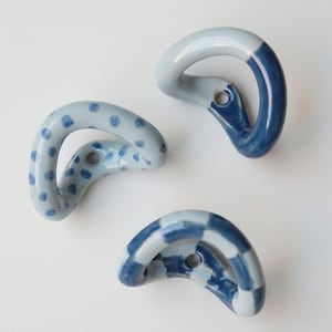 Puede incluir: Tres ganchos de cerámica en tonos azules. Uno tiene un patrón de lunares, otro es azul liso y el tercero tiene un diseño a cuadros. Cada gancho tiene una forma curva con un agujero para montar.