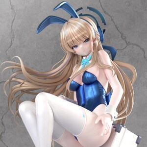 May include: A figurine of a woman in a blue bunny suit, white thigh-high stockings, and blue heels. She has long blonde hair and bunny ears. She is seated on a white suitcase with the number 77 on it.