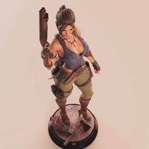 May include: A detailed figurine of a female character in a blue top, khaki pants, and brown boots, holding a rifle. The figure stands on a circular base with a hatchet and other accessories. The character has a ponytail and is wearing gloves.